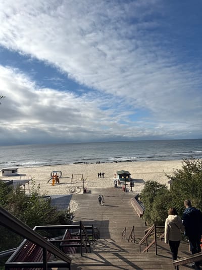 An elevated view captures the beach area of Yantarny, Russia, under a partly cloudy sky. In the immediate foreground, a wooden staircase descends towards a wide wooden boardwalk. A male in a dark jacket and a female in a light-colored hooded jacket are descending the stairs on the right. Green foliage flanks both sides of the staircase and boardwalk.

The boardwalk extends into the middle ground, where two individuals walk away from the viewer towards the beach. To the left of the boardwalk on the sand is a white rectangular structure, possibly a changing room or storage shed. To the right, a dark green hexagonal kiosk stands on the sand. Between these structures, a children's playground features a yellow and orange slide, a swing set, and a curved climbing apparatus on the light-colored sand.

In the background, the Baltic Sea meets the sandy beach. Small groups of people are dispersed along the shoreline near the water's edge. The sea exhibits light waves and white foam near the shore. The sky is predominantly covered with white and grey clouds, with patches of blue sky visible, notably a diagonal clearing in the upper center.