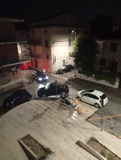 An aerial, nighttime, wide-angle shot captures a street scene in Civitanova Marche, Italy. The wet pavement reflects the light from street lamps and vehicle headlights. Several cars are parked or driving on the street. A black Audi SUV with its headlights on is driving away from the camera towards the upper right of the frame. To its left, a black hatchback is parked. In the lower left of the frame, a black Fiat 500 hatchback is parked. In the center of the frame, a silver scooter is parked. To the right of the scooter, a white Citroën hatchback is parked. Further up the street, another black Audi SUV is visible. Buildings line the street on both sides, with balconies and windows illuminated from within. Trees and streetlights are also visible. The ground is paved with large light-colored tiles and some asphalt.