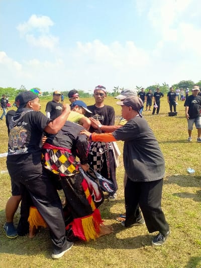 The scene depicts an outdoor gathering on a dry, grassy field in Celep, Indonesia, under a clear blue sky with scattered white clouds. The primary focus is a cluster of approximately six to seven adult males engaged in a dynamic interaction in the foreground.
At the center, one individual is dressed in black clothing, featuring a vibrant red, yellow, and black checkered fabric wrapped around the waist, accented with red and yellow fringed lower leg coverings. This individual is barefoot and appears to be actively moving or being supported. A white-beaded, red-accented object, resembling part of a headdress or an ornamental piece, is visible near their left side.
Surrounding and interacting with this central figure are several men. To the immediate left, a man in a black T-shirt with a graphic design (possibly a tiger) and a black baseball cap firmly grips the central figure's upper body. To the right, another man in a black T-shirt with visible orange long sleeves underneath, and a grey baseball cap, holds the central figure's right arm/shoulder area with both hands, seemingly providing support or guidance. Behind the central figure, a man in a black T-shirt and a blue baseball cap leans into the group, extending an arm. Further back within this central group, a man wearing a black T-shirt, a red and white patterned headband, and dark facial markings observes the interaction, with a white checkered cloth draped over his shoulder. Another man in a black T-shirt and light grey baseball cap stands slightly to the right, also observing.
In the mid-ground and background, numerous other adult males, predominantly in black T-shirts and dark pants or shorts, are dispersed across the field, some observing and others standing casually. A black bucket is visible on the grass in the mid-ground, and a white goalpost frame is discernible in the distant background. A faint white line is visible on the grass in the foreground. The scene is brightly illuminated by direct sunlight, casting distinct shadows.