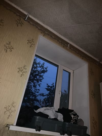 The image shows an indoor scene with a window overlooking trees and the sky at dusk. On the windowsill, a dark green suitcase with wheels is piled with various clothing items, including black, white, and striped garments. The walls are covered in a yellow wallpaper with a repeating ornate pattern. The ceiling appears to be textured and is partially obscured by a white trim. There are no people visible in the image. The overall impression is one of a slightly disheveled domestic space.