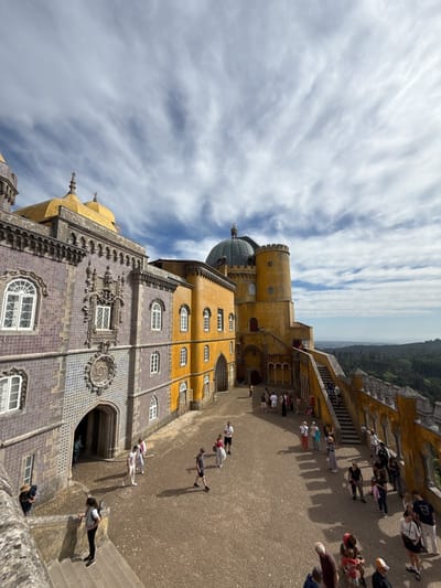 The image captures a scene at the Pena Palace in Sintra, Portugal. The foreground shows a courtyard paved with cobblestones, where a number of tourists are walking and standing. The courtyard is surrounded by architectural structures of the palace. On the left is a building with blue and gray tiles and ornate carvings. Adjacent to it is a yellow building with white-framed windows. A staircase leading to an upper level is visible along the right edge of the courtyard, with people ascending and descending. In the background, beyond the palace walls, lies a view of a forested landscape under a cloudy sky.