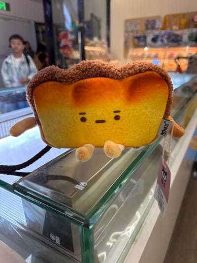A plush toy, designed as a stylized slice of toast or bread, rests centrally on a clear glass display case within an indoor commercial establishment in Guangming District, China. The toy features a textured, dark brown, crust-like top, a smooth yellowish-orange body, two small black circular eyes, and a horizontal black line mouth. Four small, light brown, stubby appendages are visible at its base. A black strap is attached to the toy's left side, and a white product tag, marked with Chinese characters "呜啦啦" and a barcode, hangs from its right side, resting against the glass. The background is blurred, revealing additional glass display cases holding unspecified merchandise under artificial lighting. To the left, a blurred reflection of a young individual, characterized by short hair, a light-colored patterned shirt, and a green lanyard, is visible in a reflective surface.