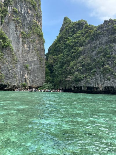 The image shows a body of clear turquoise water with a boat docked along a rocky coastline. Towering limestone cliffs covered in lush green vegetation rise steeply from the water's edge on both sides of the frame, creating a narrow passage. Several longtail boats, a common type of vessel in Thailand, are moored close to the shore, with people visible on board and disembarking. The water is calm and reflects the light, revealing the shallow sandy bottom in some areas. The sky above is a clear blue with a few wispy clouds. This scene is characteristic of the stunning natural beauty found in areas like Haad Yao, Thailand, known for its dramatic karst formations and pristine waters.
