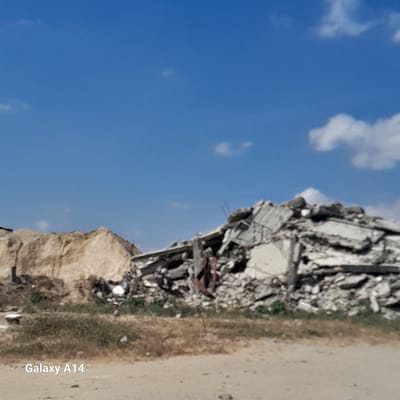 The image shows the aftermath of a destroyed building in Al-Qarara, Palestinian Territories. Rubble is piled high, next to a sandy area with sparse vegetation. The sky is blue with scattered clouds. "Galaxy A14" is visible in the lower right corner, indicating the phone model used to take the picture. There are no people visible in the frame. The scene suggests a recent demolition or bombing.