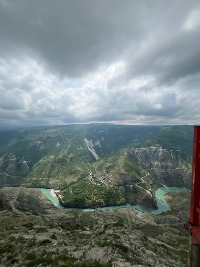 A high-angle, eye-level view captures the Sulak Canyon near the village of Dubki in Russia. A river with a turquoise hue snakes through the canyon floor, flanked by steep, rocky cliffs and verdant vegetation. The sky is predominantly overcast, with varying shades of gray clouds casting a diffuse light over the landscape. A portion of a red structure, possibly part of an aircraft or vantage point, is visible in the upper right corner of the frame.