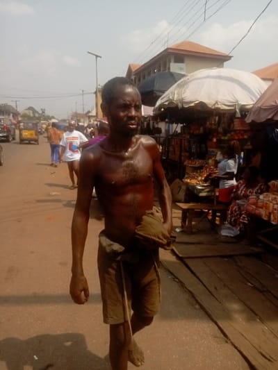 A barefoot, shirtless man walks along a street lined with vendor stalls and pedestrians. He is dark-skinned, sweaty, and appears thin, with a cloth tied around his waist and another clutched in his hand. The backdrop shows a bustling outdoor market under a partly cloudy sky, with various stalls selling goods under umbrellas and canopies. A yellow taxi-like vehicle can be seen further down the street among other cars.