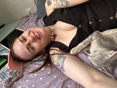 A woman with long brown hair and a black top lies on a polka-dot patterned bed. Her head rests near a pink phone case with a braided strap and a tablet device. She is smiling. A cat is sleeping next to her on the bed. Tattoos are visible on both arms and chest. A beaded necklace is visible on her chest. An electrical outlet is visible on the wall behind the bed. The location is inside an apartment. There is no information in the image that identifies the location as Novoaleksandrovka, Russia.