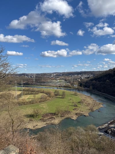 This forensic description details a panoramic landscape captured from an elevated viewpoint overlooking the Ruhr River valley in Hattingen, Germany, likely from the vicinity of Isenburg.

The sky occupies the upper half of the frame, presenting a clear, bright blue expanse punctuated by numerous scattered, fluffy white cumulus clouds. Sunlight is strong, indicating daytime.

In the immediate foreground, to the bottom left, are the bare, intertwining branches of deciduous trees, partially obscuring a rough, light-colored stone structure, possibly part of a railing or wall. The ground beneath these trees is covered in dry leaves and sparse undergrowth.

The midground features a prominent, S-shaped meander of the Ruhr River, flowing from the left and curving around a large, irregularly shaped peninsula of land. The river water exhibits a murky blue-green hue, with some ripple patterns visible. This peninsula is predominantly covered in vibrant green grass, suggesting early spring growth or dormant winter grass. Scattered across this grassy area are several leafless deciduous trees. Two large, lattice-structured electrical transmission towers (pylons) stand on the peninsula, supporting power lines that stretch across the valley.

Beyond the river, on the opposing bank, a substantial electrical substation or switchyard is clearly visible. It comprises an intricate network of metal structures, insulators, and high-voltage lines. Further across, a dense residential area with numerous buildings and houses extends into the background, sprawling across gently rolling, mostly deforested hills. More power lines and pylons are discernible within this distant urban landscape.

The right side of the image reveals a forested hillside descending towards the river. At the very bottom right, a segment of a road or pathway runs parallel to the river, with a multi-story building featuring a dark roof situated beside it.

No human figures, animals, or discernible actions are visible within the scene. The leafless state of most trees suggests a late winter or early spring setting.