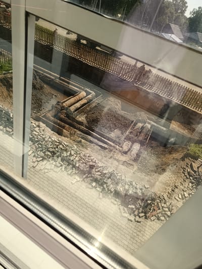 The image depicts an outdoor construction site, likely in Birobidzhan, Russia, viewed through a window, which creates reflections and distortions. The central focus is a trench where large, rusty pipes are being laid or replaced. Piles of dirt and excavated material surround the trench. On the left side of the trench, a large quantity of paving stones or bricks are stacked, suggesting recent demolition or preparation for repaving.

In the background, beyond a wooden fence, a paved area is visible with a few people and vehicles. One person, appearing to be a construction worker, is standing near the fence. Further in the distance, trees and several buildings with angular roofs are present, characteristic of a suburban or rural setting. The overall scene suggests ongoing infrastructure work in a populated area.
