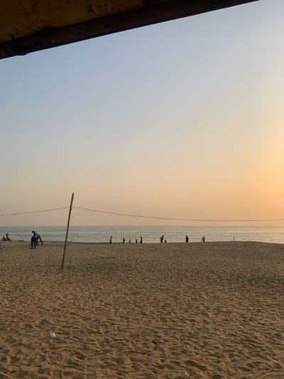 This image depicts a serene beach scene at what appears to be either sunrise or sunset, characterized by the warm, muted orange and grey hues of the sky. The horizon line neatly divides the sky from the ocean and beach.
In the upper portion of the frame, the sky transitions from a soft, pale blue-grey on the left to a warmer, peach-orange glow towards the right, indicative of a low sun. A dark, angular architectural element, possibly a roof or overhang, frames the top-left corner, suggesting the photo was taken from beneath a structure.
A thin, calm stretch of ocean water runs horizontally across the mid-ground. Along the water's edge, several distant, silhouetted figures of people are scattered, some appearing to be standing in the shallow water, others walking on the wet sand. Their forms are indistinct, adding to the tranquil atmosphere.
The lower half of the image is dominated by the textured, light brown sand of the beach, showing ripples and footprints. A prominent wooden pole, resembling a net post for a beach sport, stands upright in the lower-middle portion of the sand. A thin line, possibly a net or rope, extends horizontally from this pole towards the right, roughly aligning with the horizon. Two figures are more clearly visible near the base of this pole, one appearing to be kneeling or bending over, possibly engaged in setting up or adjusting equipment. Further into the foreground on the sand, small pieces of litter, including a crumpled white object, are visible.
The overall scene conveys a sense of peaceful leisure at a coastal location, with people enjoying the end or beginning of the day on the beach. No visible text is present in the image. Given the provided context, this scene is set in Goderich, Sierra Leone.