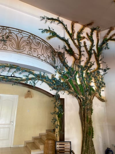 This is a medium-shot, eye-level, interior photograph of a decorative tree installation on a wall. The tree, which appears to be made of artificial materials, is brown and green, with many branches and leaves. It extends from the floor up to the ceiling. The tree is located in a building with white walls and a beige-yellow section. To the left of the tree, there is a white door with a silver handle and a beige-yellow wall with a light fixture made of wicker hanging from it. There are also stairs with beige steps and a woven basket at the bottom. A dark brown wooden chair with a slatted seat and back is visible to the right of the stairs. In the upper left portion of the image, a gold ornate railing is visible. The overall impression is of an indoor decorative feature designed to mimic a natural element within an architectural space, likely a hotel lobby or a restaurant in Kyrenia, Northern Cyprus.