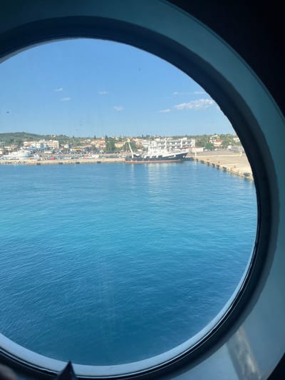 A view through a circular porthole captures a bright, sunny day in the Municipal Unit of Kastro - Kyllini, Greece. The foreground is dominated by the deep blue, slightly rippled surface of the sea, reflecting the clear sky above. Beyond the water, a harbor is visible, lined with buildings of varying heights and colors, suggesting a coastal town. Several boats are docked, including a prominent white and black vessel with the name "ALEXANDROS" clearly visible. A concrete pier extends into the water, fitted with black fenders. The background shows a gentle slope of land covered with greenery and more buildings, indicating the presence of a settlement. The sky is a vibrant blue with a few wispy white clouds. The porthole's dark rim frames the scene.