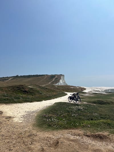 The image depicts a scenic view of the Seven Sisters cliffs in Seaford, United Kingdom, on a clear day. In the foreground, a dirt path leads to a white gravel path that winds through lush green grass dotted with wildflowers. Two bicycles are parked on the gravel path, facing towards the sea. In the middle ground, a lone figure stands near the edge of the cliff, looking out at the ocean. The iconic white chalk cliffs of the Seven Sisters stretch across the horizon, with walking paths visible on their slopes. The vast expanse of the blue sky dominates the upper portion of the image.