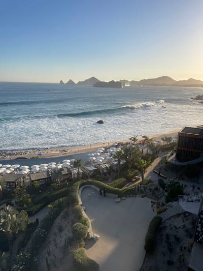 This aerial view captures a coastline in Cabo San Lucas, Mexico, during daylight. The ocean is to the left, with a large cruise ship visible on the horizon, and the beach is on the right, crowded with numerous white umbrellas and lounge chairs. A resort or hotel building is situated directly along the beach, showing a stark transition from developed land to the natural environment. A paved walkway meanders along the shoreline, bordered by manicured gardens and vegetation. In the distance, a range of mountains outlines the horizon. The sky is clear, contributing to a bright and sunny atmosphere. People are visible on the beach, suggesting active use of the resort's facilities.