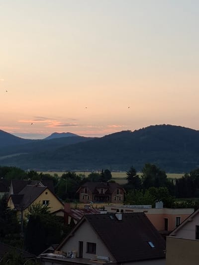 This is a photograph taken from an elevated perspective, likely a residential building, in Ilava, Slovakia, during twilight or dawn. The sky displays a gradient of soft colors, transitioning from a pale blue at the top to a warm peach and pink near the horizon, with scattered, wispy clouds. Several small, dark birds are visible in flight against the sky.
In the middle ground, a range of dark, forested hills dominates the landscape. The silhouette of a prominent, rounded mountain with a distinctive peak stands out against the lighter sky in the distance. Below the hills, a rural or suburban area is visible, featuring fields of yellow and green crops.
The foreground of the image showcases a cluster of houses with terracotta-tiled roofs. One prominent yellow house with a dark roof is visible on the left, nestled amongst trees. Further to the right, other houses with varying rooflines and architectural styles are present, some with visible chimneys and windows. The overall impression is a peaceful, serene scene of a residential area at the edge of rolling hills, bathed in the soft light of a transitional sky.