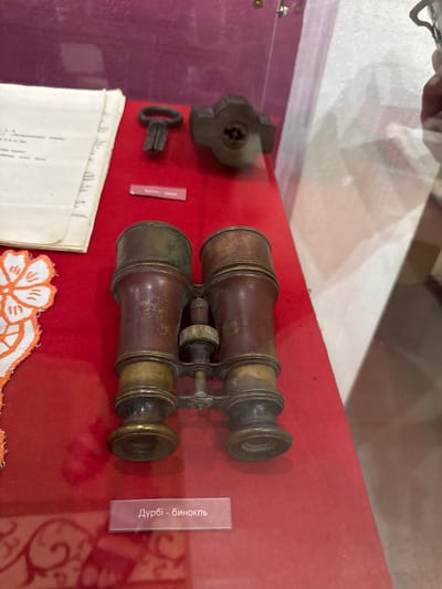 In a museum display case at the Fort-Shevchenko Museum in Fort-Shevchenko, Kazakhstan, antique binoculars rest on a red velvet surface. The binoculars are made of brass and have a worn, antique appearance. To the left of the binoculars is a stack of papers and a small embroidered white fabric with orange floral designs. Above the binoculars and to the left, there is an old key and a metal object that appears to be part of a lock. A pink label below the binoculars reads "Дурбі - бинокль" which translates to "Binoculars." The background is a deep magenta color.