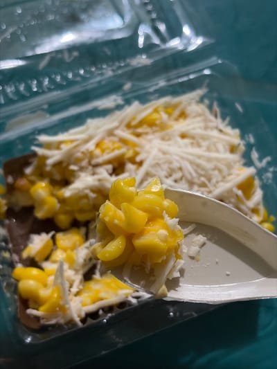 An overhead close-up shot shows a plastic container of corn kernels mixed with shredded cheese. A white plastic spoon is lifted, carrying a scoop of the corn and cheese mixture. The corn is bright yellow, and the cheese is finely shredded and white. The container is clear plastic, and the background is a blurred teal color, possibly a table. There is no indication of the city of Tupah, Indonesia, or any other specific location.