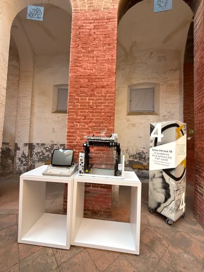 In the interior courtyard of the Old Factory of the Cooperativa Agrària in Mataró, Spain, a display features vintage computing equipment and informational signage. The scene is dominated by exposed red brickwork and pale, textured walls, suggesting an industrial or historical setting.

On a white modular shelving unit to the left, an early model personal computer with a monochrome CRT monitor and keyboard is positioned. Next to it, on another white shelf, is a glass enclosure containing what appears to be a printer or a scanner, along with electronic components.

To the right of the display, a tall, wheeled informational kiosk stands. It is decorated with graphic designs and the number "4" prominently displayed. Text on the kiosk, in Catalan, reads "Zona intrusa 19 El problema és la pintura #3 Fem soroll" followed by dates and curator information.

The environment is characterized by arched doorways and the weathered textures of the walls and flooring, which is tiled with reddish-brown terracotta. No people are visible in the image. The lighting suggests it is daytime, with light filtering in from an unseen source.