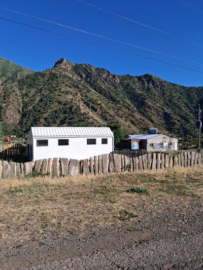 The image, taken in Shatin, Armenia, depicts a mountainside scene under a clear, bright blue sky, suggesting daytime and fair weather. The primary subjects are two small buildings situated on a steep, verdant mountain slope. One building is a modern, white, rectangular structure with three black rectangular windows. Adjacent to it is an older, multi-colored building with a blue-tiled roof and several windows, appearing to be in a state of disrepair or undergoing renovation.
A pathway made of rough-hewn wooden logs ascends the slope alongside the buildings, bordered by dry, golden grass and loose gravel. In the background, the rugged mountainside is covered in dense green foliage and rocky outcrops. Power lines stretch diagonally across the upper left portion of the image, a common sight in mountainous regions.
There is no visible text in the image. The scene conveys a sense of remote, rugged beauty and a functional, perhaps slightly rustic, human presence in a natural environment. The presence of the buildings suggests habitation or a point of interest within the mountainous landscape. The lighting indicates it is likely mid-morning or afternoon.