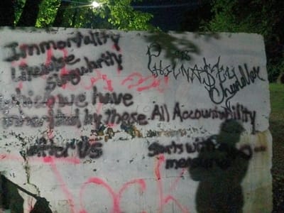 An outdoor scene at night or dusk features a prominent concrete wall covered in graffiti. Lush green foliage, likely trees or bushes, is visible in the upper background, illuminated by an unseen light source in the top left. A faint, dark shadow of a person, presumably the photographer, is cast on the lower right portion of the wall.

The wall is extensively covered with spray-painted text and symbols in black and red. On the left side, several lines of black text are visible, including "Immortality", "Life expectancy", "Singularity", "these we have", "inherited", and "murdered by these", followed by "that matter US" near the bottom. These phrases are interspersed with red spray paint that is mostly abstract marks but also underlines some words.

On the right side, there is a distinct tag "P 918" and a stylized signature "NASTY Chandler". Below this, in black spray paint, are the phrases "All Accountability" and "Starts with me", though the word "me" is largely obscured by the human shadow. The overall impression is one of a public space used for layered, expressive, and possibly political or philosophical statements through graffiti.