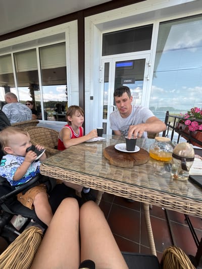 The image depicts an outdoor seating area at a cafe, likely in Akhlebaikha (Kovriginskii S/S), Russia. A man in a grey t-shirt is seated at a wicker table, reaching for a black cup. On the table, there is a wooden serving board, a glass teapot with orange liquid, and a baby bottle. A young boy in a red and white tank top is also seated at the table, eating. A baby in a stroller is to the left of the frame, holding a black cup to its mouth. In the background, large windows with blinds suggest an indoor space, and a sign on the door reads "ОФИС" (Office). The overall environment appears to be a casual dining setting, possibly by a body of water, given the hazy landscape visible through the windows.
