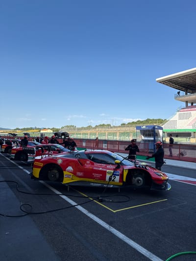 The image captures a sunny day at the Mugello Circuit, a renowned racetrack located in Scarperia, Italy. In the foreground, several red and yellow Ferrari race cars are lined up on the asphalt track, some with their hoods open and tires exposed, suggesting preparation or maintenance. Pit crew members, dressed in dark racing suits and helmets, are visible around the cars, engaged in various tasks. To the right, a section of the grandstands with white seats is visible, overlooking the track. Beyond the track, there are green hills and trees, characteristic of the Tuscan landscape surrounding Scarperia. The sky is a clear, vibrant blue, with a few wispy clouds.