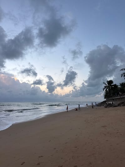 An overcast sky looms over a beach scene in Ban Bang Niang, Thailand. The sky is a muted blue and gray, with large, billowy clouds dominating the upper portion of the frame. In the distance, the ocean is calm, with gentle waves lapping at the shore. The beach itself is wide and sandy, with footprints scattered across the surface. A few people are visible on the beach, some standing and others sitting. To the right, palm trees line the shore, their fronds silhouetted against the cloudy sky. A low stone wall runs along the edge of the beach, separating it from what appears to be vegetation behind the trees. The overall impression is one of a quiet, serene beach at dusk or dawn.