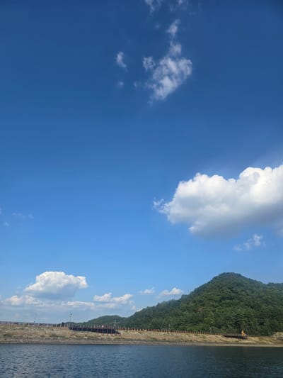 This media file is an image of a body of water with a forested mountainside in the background. The sky is a clear blue with scattered white clouds. A paved walkway with a railing runs along the edge of the water and up the side of the mountain. There is a yellow construction vehicle visible on the walkway near the top of the mountain. The water is calm and appears to be reflecting the blue sky. The overall scene evokes a sense of natural beauty and tranquility. The time of day appears to be daytime, likely late morning or early afternoon, given the bright sunlight. The weather is clear and sunny. The location is Gumi-si, South Korea. No people are visible in the image. No text is visible in the image.