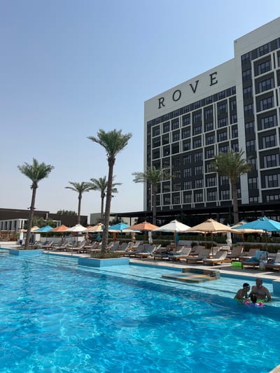 This is a photograph taken outdoors at what appears to be a resort or hotel with a swimming pool and a tall building. The scene is dominated by a large, bright blue swimming pool. In the pool, two people are visible, appearing to be a father and son. The father is standing and looking towards the son, who is younger and wearing goggles. They are interacting with some colorful pool toys.

Around the pool, there are numerous lounge chairs, many of which are empty, suggesting a leisurely daytime activity. Several palm trees are scattered throughout the scene, their fronds reaching towards a clear, light blue sky. Behind the palm trees and lounge chairs, a large, modern building stands. The building has a dark facade with a grid-like pattern of windows. The word "ROVE" is prominently displayed in large, dark lettering on the side of the building.

The lighting suggests it is daytime, likely sunny, given the bright blue of the pool water and the clear sky. There are umbrellas providing shade over some of the seating areas. The overall mood seems relaxed and vacation-like. The image is oriented vertically, with the building and palm trees on the left side and the pool and people on the right.