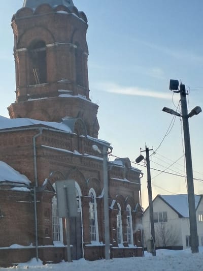 The image captures a winter scene featuring a large, ornate brick building, likely a church or a similar historic structure, under a bright, clear sky. The building dominates the left and central-right portions of the frame.
The main structure is constructed from reddish-brown brick, exhibiting detailed architectural elements. It features numerous arched windows with white frames, many of which have accumulated snow on their sills and ledges. The building has multiple levels and decorative brickwork along its cornices and other horizontal surfaces, also dusted with snow. To the left, a tall tower-like section rises, seemingly a bell tower, showcasing similar intricate brick patterns and arched openings. Its uppermost part is partially visible, hinting at a dome or cupola structure.
In the upper right quadrant of the image, against the blue sky, a utility pole stands prominently. It has several cross arms supporting an array of wires and two street lights. One street light is a dark, somewhat square-shaped fixture, while the other is a more common curved design. A faint, white contrail or wispy cloud stretches across the clear blue sky, extending from the upper left towards the lower right.
In the background, behind the brick building and utility pole, a smaller, white-painted building with a dark roof and windows is partially visible, also appearing to have snow on its roof. The immediate foreground and right edge of the image show indistinct, snow-covered ground or snow accumulation.
The scene appears to be set during the daytime, indicated by the bright illumination and clear sky. The presence of snow on all surfaces confirms a winter season. There are no people visible, and no specific activity is taking place. No text is discernible in the image.