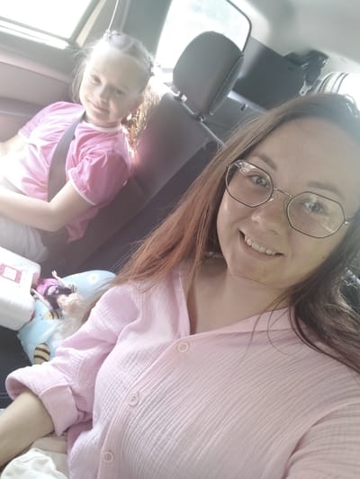 A medium shot photograph captures two individuals, a woman and a young girl, inside a car in what appears to be a suburban or rural setting, potentially in or near Степное Полеологово, Russia. The woman, positioned in the foreground, is wearing glasses and a pink textured blouse, and she is smiling towards the camera, holding the camera and taking a selfie. To her left and slightly behind her, a young girl with braided hair is visible, wearing a pink t-shirt and a seatbelt. The girl is looking slightly away from the camera, also with a gentle smile.
The interior of the car is dimly lit, with light streaming in from a window on the left side of the frame. The background shows car seats, headrests, and a glimpse of the outdoors through the windows. A white plastic container with a pink label and a doll are on a pillow between the woman and the girl. The overall atmosphere suggests a casual moment during a car journey.