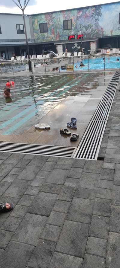 This image captures a scene at an outdoor swimming pool complex in Tulskiy, Russia. The main subjects are people enjoying the water and various objects left by pool-goers. The setting is a tiled area adjacent to a swimming pool, with a building featuring a vibrant mural on its side.
Several individuals are visible in the pool, some submerged and others with their heads above water. One person is wearing an orange flotation device. On the wet tiled ground near the pool, there are several pairs of footwear, including flip-flops and sandals, indicating recent use. A white object, possibly a towel or another piece of clothing, is also present on the ground.
The building in the background is adorned with a colorful mural depicting tropical foliage and mountains. Above the entrance, a digital display shows "18:22:0", likely indicating the time or temperature. The word "САУНА" (SAUNA in Russian) is also visible on the building. A yellow sign with Russian text, which appears to be a warning or rule, is posted near the pool.
The ground is wet and reflects the sky and surrounding structures, suggesting it might have recently rained or that the pool area is actively being used and watered. The lighting and reflections indicate it is likely daytime, possibly late afternoon given the time displayed. The overall atmosphere appears to be one of leisure and relaxation at a recreational facility.