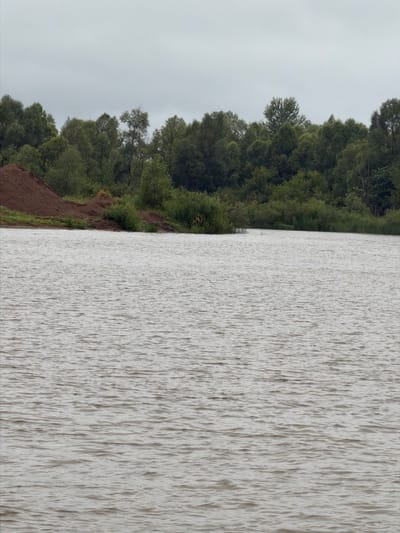 The image depicts a body of water with a shoreline lined by trees and a pile of dirt. The sky is overcast. The water has small ripples on its surface. The trees are a mix of deciduous and coniferous species, with dense foliage. The dirt pile is reddish-brown and appears to be construction material. There are no people or animals visible in the image. The location is Azyakul', Russia.