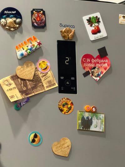 The image is a close-up, eye-level shot of a gray refrigerator door adorned with various magnets and small decorative items. The lighting is even and diffused, suggesting indoor ambient light. No specific city or location information is present in the image itself, nor can it be deduced from the visual content.
On the top left, a round magnet features the word "Абхазия" in stylized white lettering over a dark blue background with images of shells and stones. To its right, a rectangular magnet depicts a tiger's face with the word "Виктория" above it. Further right, a magnet shows a red Christmas stocking with a green bird and berries, with text below that is partially visible. Another magnet to the far right appears to be a small postcard with a picture of a tiger.
Below these, a colorful, three-dimensional magnet spells out "АХАНА" with seashells and a red starfish. A large, black rectangular panel with digital readouts and buttons (displaying "2°C" and text like "SUPER COOL," "SET TEMP," and power icons) is visible on the left side.
A heart-shaped wooden magnet engraved with "08.07.2023" and floral patterns rests on top of a 500-ruble banknote. Adjacent to this is a round magnet with a laughing emoji face. A round, colorful magnet with a frog character and text that may say "С Новым годом!" is positioned below the black panel.
To the right of the banknote, a large red heart-shaped magnet features the text "С 14 февраля Люблю тебя!!!" and a small photograph of a couple kissing. Two small black square magnets are attached to the top right of the heart magnet.
Below the frog magnet, a golden framed photograph shows a wedding party consisting of a couple in the center, a man in a suit and red tie to the left, and a woman in a light pink dress to the right. Two small, round, colorful bottle cap magnets are placed above the framed photo.
On the lower left, a round magnet depicts a yellow smiley face wearing black sunglasses. Another heart-shaped wooden magnet, similar to the one on the banknote, is located in the lower right, also engraved with text that includes "08.07.2023."
The overall impression is that of a personal collection of souvenirs and mementos on a refrigerator door.