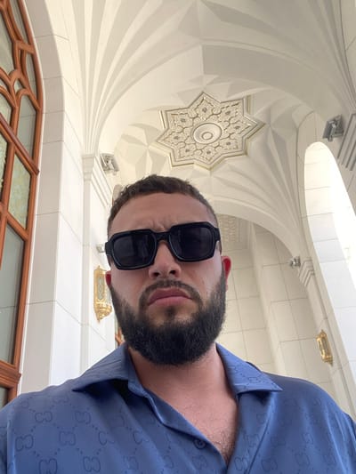 A man with a beard and sunglasses is captured in a selfie, looking directly at the camera. He is wearing a blue collared shirt with a repeating pattern of interlocking "G" shapes. The background features the ornate architecture of the Shali Grand Mosque in Shali, Russia. The mosque's interior is characterized by white walls and intricate geometric patterns on the ceiling, with sunlight filtering through a large wooden window.