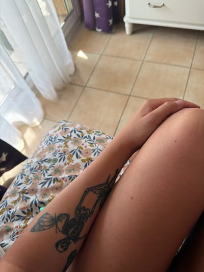 The media file shows a close-up view of a person's arm and leg, with a tattoo of a skeleton and a butterfly visible on the arm. The person is wearing a floral patterned dress or skirt. The background consists of a tiled floor and a white cabinet with a silver handle. To the left of the cabinet, there are stacked purple and white objects with star patterns. Sunlight is visible, casting shadows and suggesting it is daytime. The scene appears to be indoors, possibly a bedroom or dressing area, given the furniture and tiled floor. There is no visible text in the image. The context provided suggests the location is Sainte-Marie-la-Mer, France. The image is a still shot, and no specific activity or event is taking place. The focus is on the person's arm and tattoo.