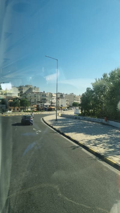 The image depicts a street scene in Larissa, Greece, viewed from inside a vehicle. In the foreground, an asphalt road curves to the right, with white lane markings visible. A dark blue car is traveling on the road. To the right of the road is a sidewalk paved with square tiles, bordered by a curb with yellow and black stripes. A lamppost stands on the sidewalk.
Beyond the sidewalk, a line of buildings forms the urban landscape. These buildings are multi-story structures with light-colored facades, exhibiting a common architectural style. Some buildings have balconies and windows. Trees are visible interspersed with the buildings and on the right side of the image, a large tree casts shadows on the sidewalk.
In the distance, the sky is clear and blue with a few wispy clouds. There is some graffiti on the wall behind the sidewalk. The overall impression is of a sunny day in an urban environment.