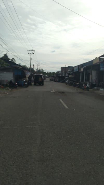 The image depicts a street scene in Deah Pangwa, Indonesia, under an overcast sky. The foreground is dominated by a grey asphalt road with a white dashed line marking the center. On the left side of the road, there are several parked motorcycles and a dark-colored SUV with its rear hatch open. Adjacent to the motorcycles are small shops or market stalls with open fronts. On the right side of the road, a row of similar market stalls or shops with dark roofs stretches into the distance. More motorcycles are parked in front of these establishments. In the background, power lines crisscross the sky, supported by utility poles. Trees and some low-lying structures are visible on the far left. The overall impression is that of a bustling local market or commercial area along a road.