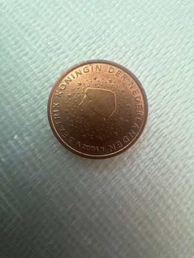 The image shows a close-up, overhead view of a Dutch 5 Euro cent coin lying on a white, textured surface, likely fabric. The coin is copper-colored and features a profile of Queen Beatrix of the Netherlands facing left, surrounded by stars. The inscription on the coin reads "KONINGIN DER NEDERLANDEN" (Queen of the Netherlands) and "BEATRIX A 2004". The coin is positioned slightly off-center in the frame. The background is blurred and appears to be a soft, possibly fuzzy material. There is no indication of the city of Schkopau, Germany, in this image.