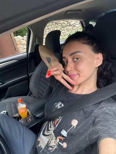 A woman with dark hair is sitting in the passenger seat of a car. She is wearing a dark gray t-shirt with a graphic print and dark pants. Her right arm is resting on her lap, and she is holding up her left hand with her index and middle fingers extended in a peace sign. She is looking at the camera and smiling. A bottle of orange liquid and a can are visible in the center console of the car. The interior of the car is dark-colored. Outside the car window, a stone wall and some greenery are visible. The image appears to be taken in Manavi, Georgia.