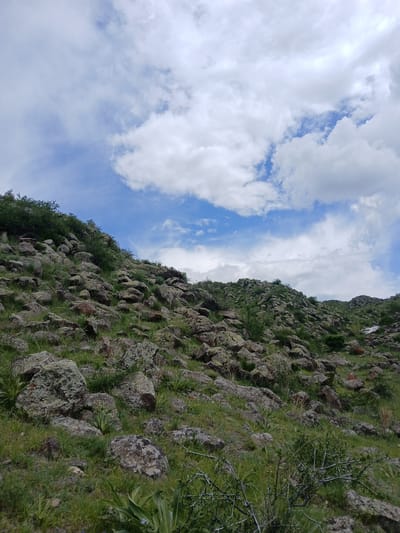 The image depicts a rocky hillside in the city of Tlik, Armenia, ascending diagonally from the lower right to the upper left. The hillside is covered in rocks of varying sizes, interspersed with patches of green grass and small shrubs. The sky above is partly cloudy, with patches of blue visible through the white clouds. There are no people visible in the scene. The focus is on the natural environment, specifically the rocky hillside and the sky.