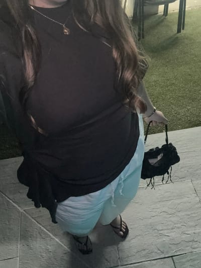 In the outdoor area of Urbanización Encinares De Alcolea, Spain, a person is captured from the waist down, holding a small black bag with fringe details. The person is wearing light blue shorts and dark flip-flops. They are standing on a tiled patio, with a patch of green artificial turf visible in the background, along with dark-colored outdoor furniture. The lighting suggests it may be evening. The person is also wearing a black top and a necklace.