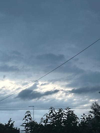 The image captures a cloudy sky over what appears to be a residential area. A thin, dark wire stretches diagonally across the upper portion of the frame, from the top left towards the bottom right. Below the wire, the sky is filled with varying shades of gray and white clouds, suggesting overcast or stormy weather. In the lower part of the image, the silhouettes of dark green foliage and tree branches are visible. A thin, horizontal antenna or mast stands among the trees on the left side, with two parallel wires running horizontally behind it. The scene is likely outdoors, potentially in a backyard or garden, given the presence of trees and the antenna. The specific location in Prokhladny, Russia cannot be determined from the visual information alone.