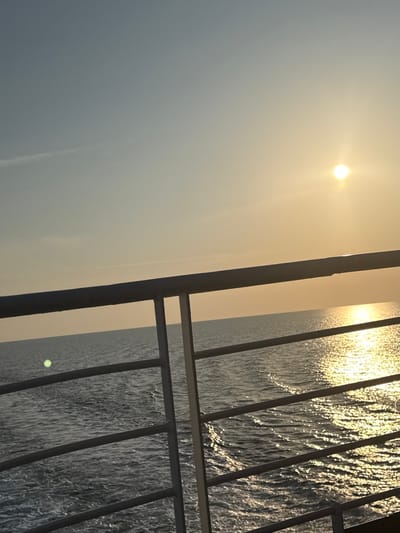 The image displays a maritime scene from the perspective of a vessel at sea, located in Kato Karioutes, Greece. The foreground features a grey metal railing, likely part of a ship's deck, positioned horizontally across the lower third of the frame. This railing consists of a thick top bar and several thinner horizontal bars supported by vertical stanchions. Beyond the railing, the midground shows the dark blue surface of the open sea with visible ripples and a wake indicating the vessel's movement. A prominent golden reflection path from the sun stretches across the water towards the viewer. The background consists of a clear sky, transitioning from a light blue-grey in the upper left to a warm yellow-orange hue near the horizon. A bright, intensely glowing sun, appearing as a circular point of light, is positioned in the upper right quadrant, close to the horizon line. A faint, thin contrail or wispy cloud is visible in the upper left sky, above the horizon. A distinct green lens flare is present on the water surface in the lower-left area of the frame. No individuals are visible. The lighting conditions suggest either sunrise or sunset.