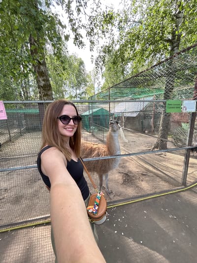 A woman is taking a selfie in an outdoor enclosure with a llama. The woman, wearing a black tank top, sunglasses, and olive green pants, is smiling at the camera. Her arm is extended, holding the camera. A small, round, brown purse with a colorful strap is visible on her hip. Behind her, a llama stands in a dirt enclosure surrounded by a chain-link fence. In the background, there are several small structures with green netting overhead, and trees are visible throughout the scene. A sign with Russian text is attached to the fence near the llama. The location is identified as Medvedevo Urban Settlement, Russia.