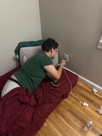 The image captures a young male lying on his side on a bed or mattress in an indoor room. He has dark, curly hair and wears glasses, a green t-shirt, and white boxer briefs with "FRUIT OF THE LOOM" visible on the waistband. His right hand is extended to a white wall outlet, where he is actively plugging in a white charging cable. In his left hand, he holds a small, black electronic device, possibly a remote control or a portable gaming device.
The setting appears to be a personal space, likely a bedroom, characterized by light grey walls and a light-toned wooden floor. A maroon comforter is draped over the lower half of his body and the bed. A light grey pillow and a dark green pillowcase are visible beneath his head.
On the wooden floor, several items are scattered: two crumpled pieces of white paper or tissue, a partially full clear plastic water bottle, and a white power adapter. A long white charging cable extends from the wall outlet towards the man. The scene depicts a casual moment, possibly at night or indoors with artificial lighting, with the man focused on connecting a device to power. The overall atmosphere is relaxed and unposed.