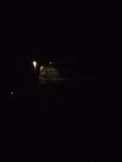 The image, taken at night in the village of Zaplavnoye, Russia, shows a dimly lit scene. In the center, a greenhouse-like structure is visible, illuminated by a light source above it. The structure appears to be made of a translucent material and has a curved roof. To the left of the greenhouse, a small, faint blue light is visible in the darkness. The rest of the image is predominantly black, indicating a low light environment with limited visibility. No people or specific actions are discernible due to the darkness.