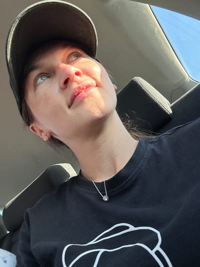 A woman is posing for a selfie inside a car. She is wearing a black baseball cap and a black sweatshirt with a white graphic design on it. She has a silver necklace on. The interior of the car is visible, including the headrest of the car seat. The background shows the sky and a portion of the car's interior. The image was not taken in Langwedel, Germany.