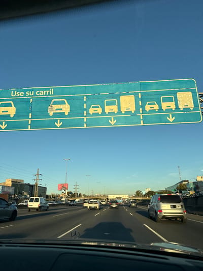 The image shows traffic on a highway in Boulogne Sur Mer, Argentina. A large blue sign indicates which vehicles are allowed in a particular lane, including buses, vans, and cars with luggage. The sign also has the Spanish phrase "Use su carril," which means "Use your lane." The traffic is moderate to heavy, with a variety of vehicles visible. The sky is clear and blue.