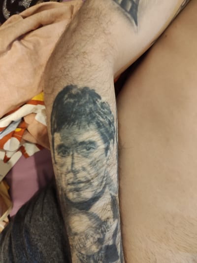 The image shows a close-up view of a person's arm with a tattoo of a face. The tattoo is a black and white portrait, and it appears to be of Al Pacino as Tony Montana from the movie Scarface. The person's arm is hairy, and the tattoo is on the forearm. The person is lying down, and there are some blankets in the background. The lighting is dim, suggesting it might be indoors. There is no visible text in the image. The location context of Lisakovsk, Kazakhstan is not visually evident in this image.